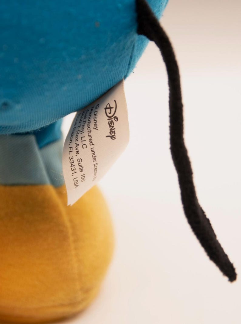 May include: A plush toy featuring a bright blue fabric section, a yellow fabric base, and a thin black tail. A white fabric tag is attached, displaying the Disney logo and text: Manufactured under license by Just Play, LLC, 4850 T-Rex Ave, Suite 100, Boca Raton, FL 33431, USA.