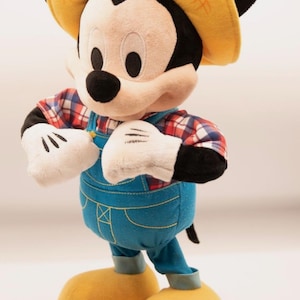 May include: A plush Mickey Mouse doll wearing a yellow straw-style hat, a red and white plaid shirt, and blue denim-colored overalls with yellow stitching. The character has white gloves, black ears, and large yellow shoes, standing in an upright pose.