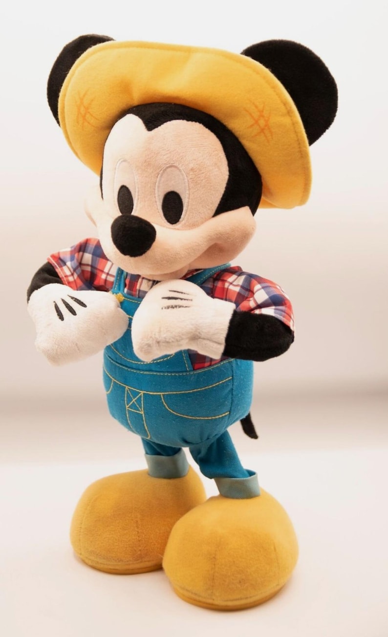 May include: A plush Mickey Mouse doll wearing a yellow straw-style hat, a red and white plaid shirt, and blue denim-colored overalls with yellow stitching. The character has white gloves, black ears, and large yellow shoes, standing in an upright pose.