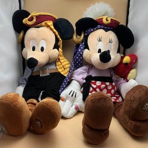 May include: Two plush pirate-themed Mickey and Minnie Mouse dolls. Mickey wears a red and gold pirate hat, gray shirt, black pants, and brown boots. Minnie wears a purple polka-dot headscarf, a red and white polka-dot skirt, and brown boots.