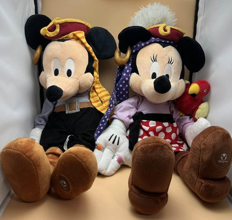 May include: Two plush pirate-themed Mickey and Minnie Mouse dolls. Mickey wears a red and gold pirate hat, gray shirt, black pants, and brown boots. Minnie wears a purple polka-dot headscarf, a red and white polka-dot skirt, and brown boots.