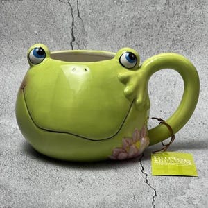 May include: A lime green ceramic mug shaped like a frog with large blue eyes, a wide smile, and a handle resembling a frog leg. A small pink flower is painted on the side. A green tag attached to the handle displays the text burton + BURTON.