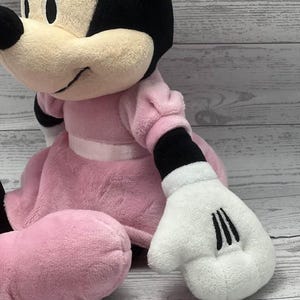 May include: A plush Minnie Mouse doll. The doll is wearing a pink dress with a white ribbon, black gloves with three black lines, and black ears. The doll has a cream-colored face and a black bow.