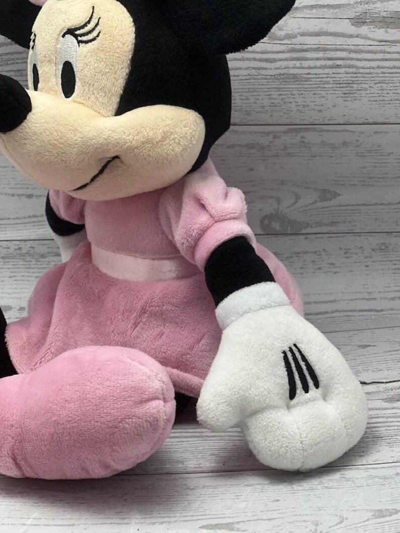 May include: A plush Minnie Mouse doll. The doll is wearing a pink dress with a white ribbon, black gloves with three black lines, and black ears. The doll has a cream-colored face and a black bow.