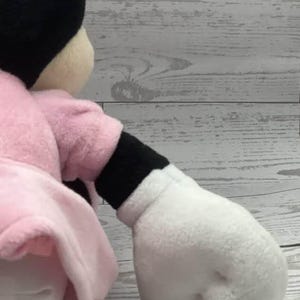 May include: A plush Minnie Mouse doll, featuring a black head with large ears, a pink dress, and white gloves. The doll's arm is extended, showcasing the white glove and black sleeve. The doll is set against a light wood-grain background.
