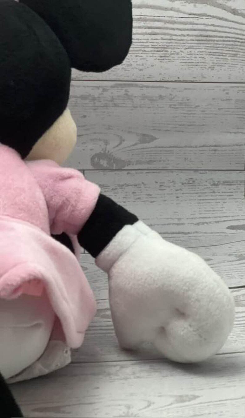May include: A plush Minnie Mouse doll, featuring a black head with large ears, a pink dress, and white gloves. The doll's arm is extended, showcasing the white glove and black sleeve. The doll is set against a light wood-grain background.