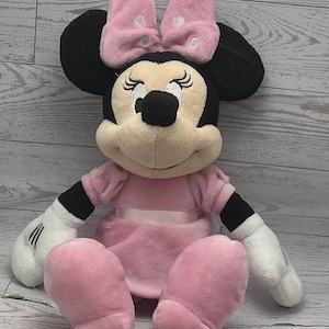 May include: A plush Minnie Mouse doll, featuring a pink dress, bow, and shoes. The doll has black ears, a black nose, and white gloves. The bow has white polka dots. The doll is sitting down.