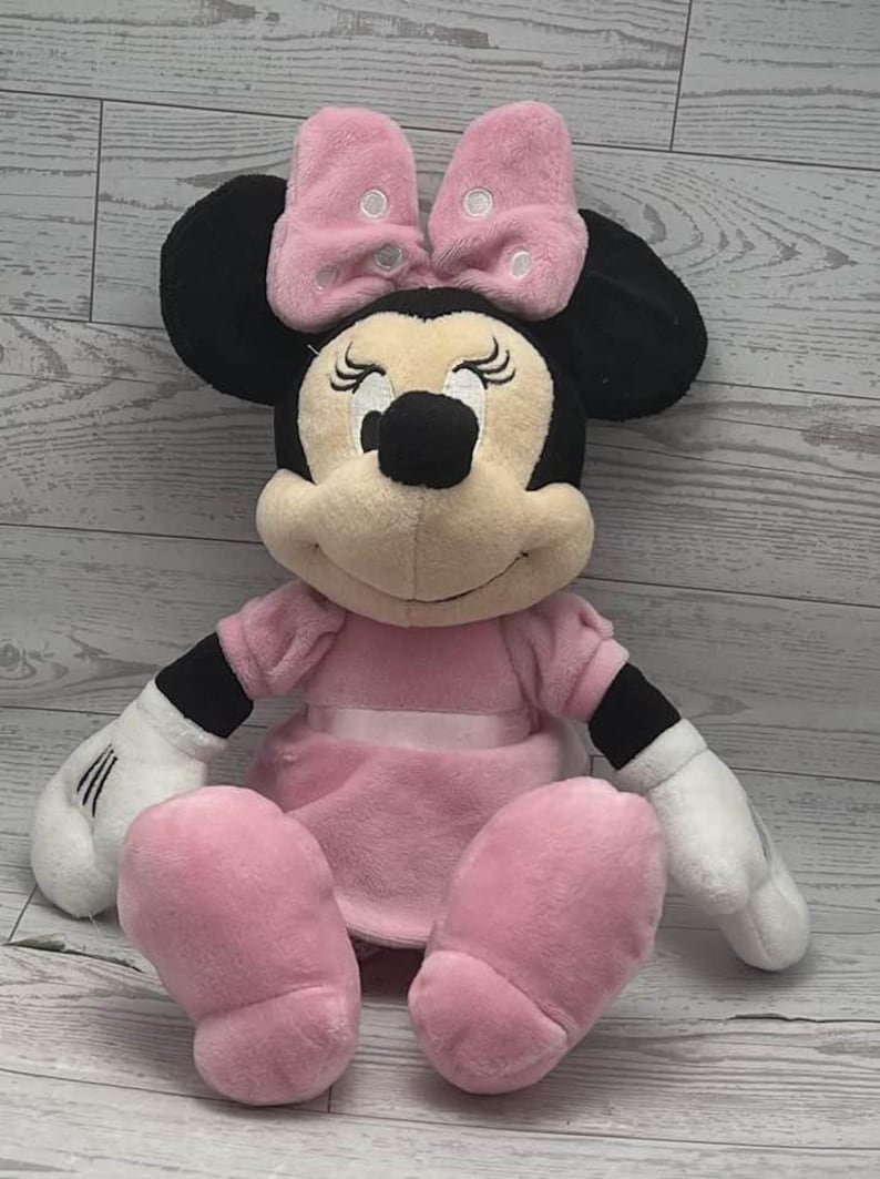May include: A plush Minnie Mouse doll, featuring a pink dress, bow, and shoes. The doll has black ears, a black nose, and white gloves. The bow has white polka dots. The doll is sitting down.