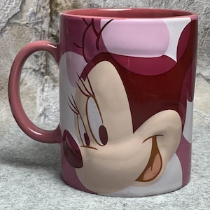 Op de afbeelding: Een keramische mok met een afbeelding van Minnie Mouse aan de buitenkant. De mok heeft een roze binnenkant en een handvat, met een kleurenpalet bestaande uit verschillende tinten roze, donkerrood en wit, met een groot karaktergezicht.