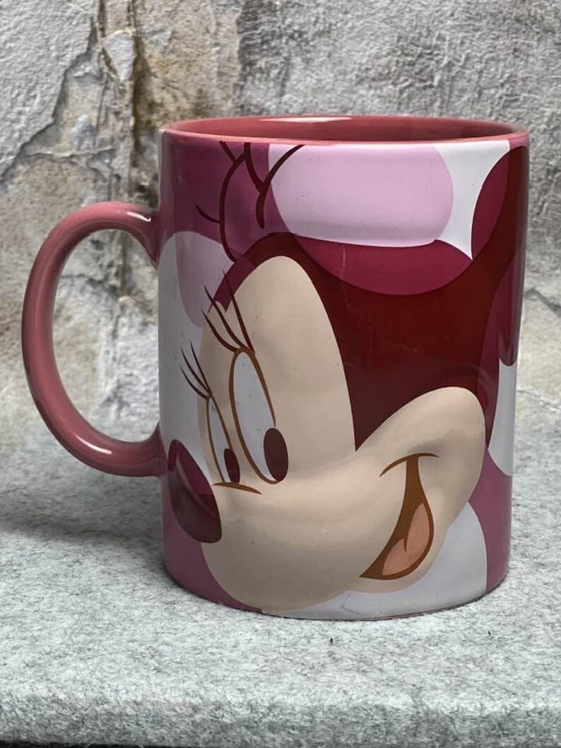 Op de afbeelding: Een keramische mok met een afbeelding van Minnie Mouse aan de buitenkant. De mok heeft een roze binnenkant en een handvat, met een kleurenpalet bestaande uit verschillende tinten roze, donkerrood en wit, met een groot karaktergezicht.