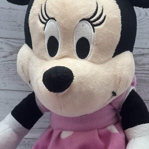 Pode incluir: Uma pel&uacute;cia da Minnie Mouse, com cabe&ccedil;a e orelhas pretas, um la&ccedil;o rosa e um vestido rosa com bolinhas brancas. A boneca tem uma apar&ecirc;ncia macia e texturizada com tra&ccedil;os faciais bordados.