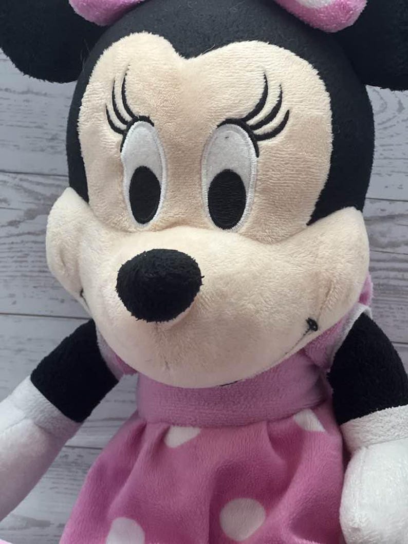 Pode incluir: Uma pel&uacute;cia da Minnie Mouse, com cabe&ccedil;a e orelhas pretas, um la&ccedil;o rosa e um vestido rosa com bolinhas brancas. A boneca tem uma apar&ecirc;ncia macia e texturizada com tra&ccedil;os faciais bordados.