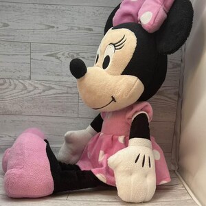 Pode incluir: Uma boneca de pel&uacute;cia Minnie Mouse sentada. A boneca tem um vestido rosa com bolinhas brancas, um la&ccedil;o combinando e luvas brancas. A boneca tem orelhas pretas e um nariz preto. O fundo &eacute; um padr&atilde;o de gr&atilde;o de madeira clara.