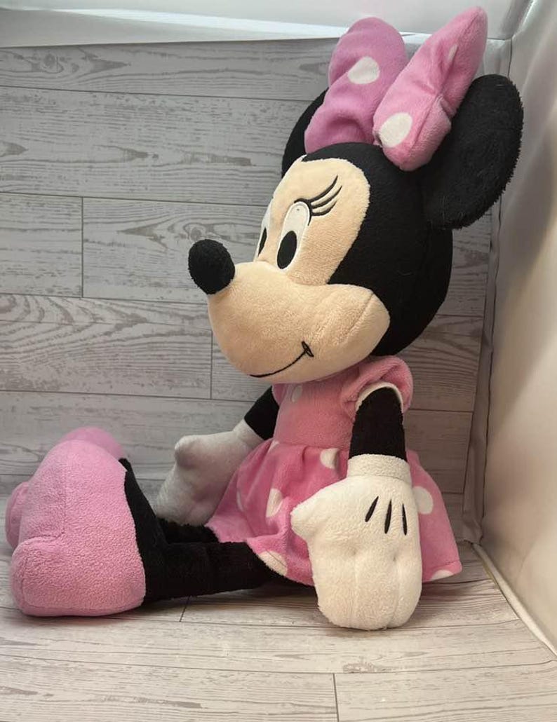 Pode incluir: Uma boneca de pel&uacute;cia Minnie Mouse sentada. A boneca tem um vestido rosa com bolinhas brancas, um la&ccedil;o combinando e luvas brancas. A boneca tem orelhas pretas e um nariz preto. O fundo &eacute; um padr&atilde;o de gr&atilde;o de madeira clara.
