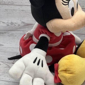 May include: A plush Minnie Mouse doll, with a red dress and white polka dots, a black head with a red bow, and white gloves with black markings. The doll has a yellow shoe and is set against a light wood background.