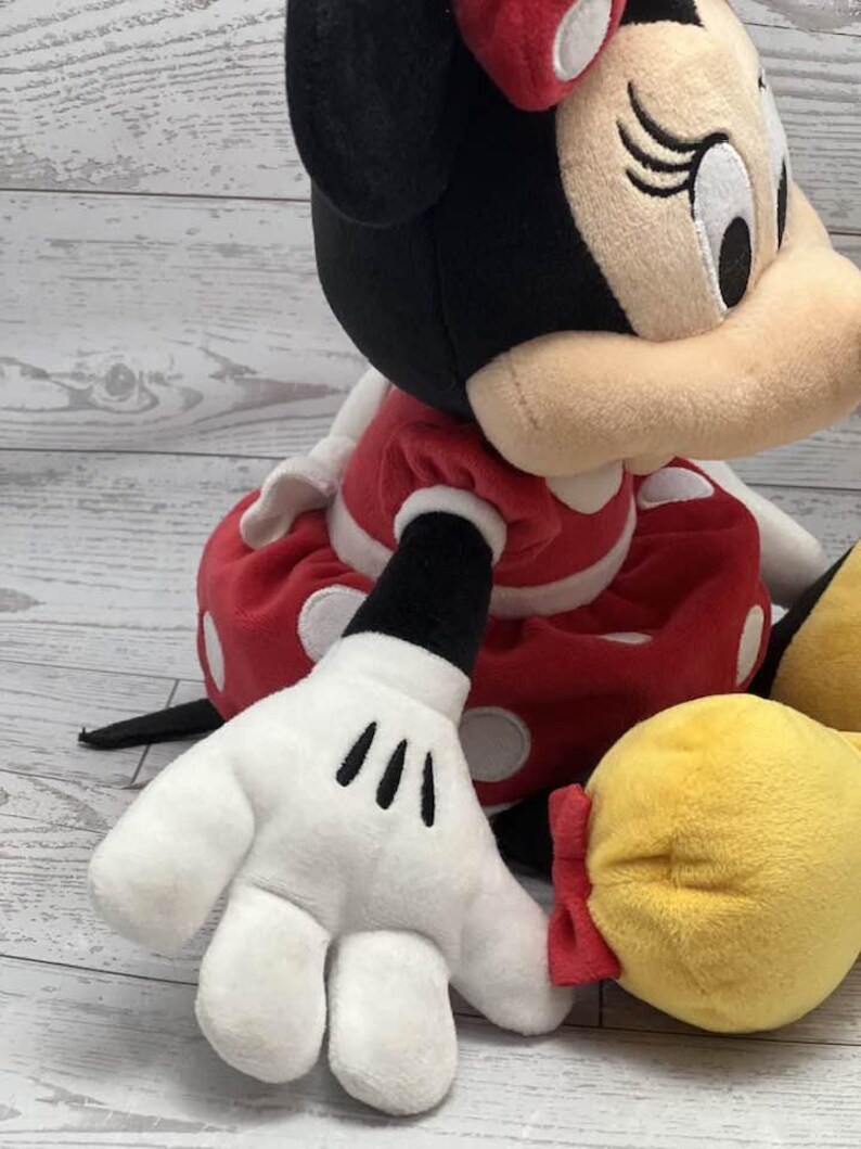 May include: A plush Minnie Mouse doll, with a red dress and white polka dots, a black head with a red bow, and white gloves with black markings. The doll has a yellow shoe and is set against a light wood background.