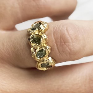 May include: A gold-colored ring with four oval-shaped, dark green gemstones. The ring has a textured, organic design, and is worn on a finger. The ring is a piece of jewelry.