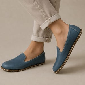 May include: A pair of blue slip-on shoes featuring a rounded toe, a low-cut topline, and a visible light-coloured stitched detail along the edge of the sole. The shoes have a smooth, matte finish and a flat, flexible construction for everyday wear.