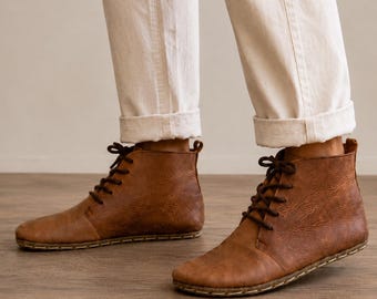 May include: A pair of ankle-high boots crafted from textured brown leather, featuring dark brown laces and visible stitching along the sole. The boots are worn with light-colored, cuffed trousers that end just above the ankle, showcasing the footwear.