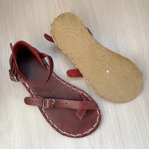 Peut inclure: Une paire de sandales en cuir rouge foncé dotées d'une semelle plate, d'une bride de cheville avec une boucle en métal et d'une bride d'orteil avec une boucle assortie. Les sandales présentent des coutures blanches visibles sur les bords.