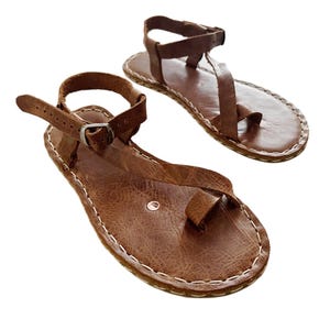 Peut inclure: Une paire de sandales en cuir marron avec un design à boucle d'orteil, une bride de cheville réglable avec une boucle en métal et des coutures blanches visibles le long du bord de la semelle. Le cuir présente une finition texturée et plate.