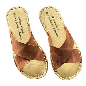 Peut inclure: Paire de sandales en cuir marron avec des lanières entrecroisées. Les sandales ont une semelle de couleur claire et des bords cousus. Le texte "MerCan Barefoot Shoes" est imprimé sur le côté de chaque sandale.