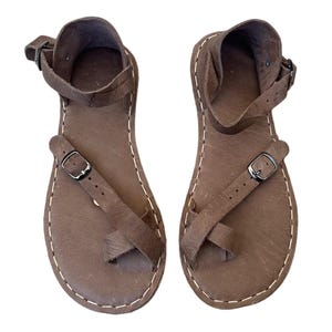Peut inclure: Une paire de sandales en cuir marron avec un design à boucle d'orteil, une bride de cheville réglable avec une boucle en métal et une bride diagonale sur le pied fixée par une plus petite boucle en métal. Les bords présentent des coutures blanches.