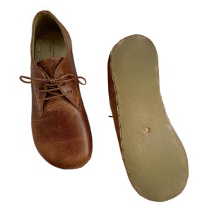 Op de afbeelding: Een paar bruine leren schoenen met veters. Eén schoen staat rechtop en de andere is omgedraaid om de zool te laten zien. De schoenen hebben een klassiek ontwerp en een getextureerde uitstraling.