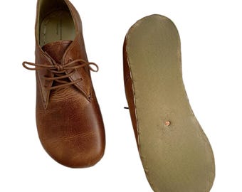 Könnte beinhalten: Ein Paar braune Lederschuhe mit Schnürsenkeln. Ein Schuh steht aufrecht, der andere ist umgedreht, um die Sohle zu zeigen. Die Schuhe haben ein klassisches Design und eine strukturierte Optik.