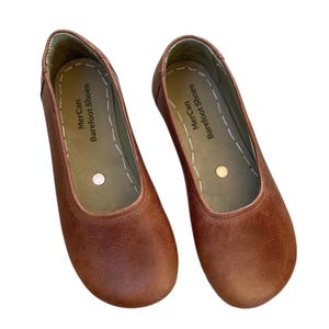 Peut inclure: Une paire de chaussures à enfiler en cuir marron avec un bout arrondi et une semelle plate. L'intérieur présente une doublure de couleur claire avec des coutures blanches sur le bord et le texte MerCan Barefoot Shoes imprimé en noir.