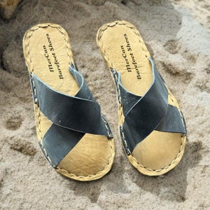 Peut inclure: Une paire de sandales à enfiler avec des brides croisées gris anthracite foncé sur une semelle intérieure beige texturée. Les bords de la semelle sont cousus avec un fil beige épais. Le texte MerCan Barefoot Shoes est imprimé au talon.