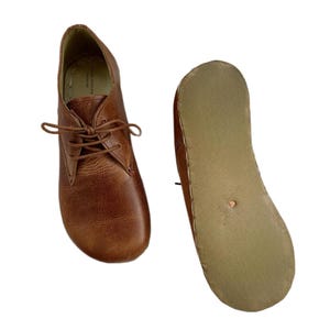 May include: A pair of tan leather shoes with a lace-up front and a rounded toe. One shoe is shown from the top, revealing the interior lining and laces, while the other is shown from the bottom, displaying a flat, light-colored sole with a small stud.