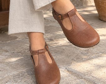 May include: A pair of brown T-strap shoes featuring a rounded toe, a closed heel, and a thin adjustable ankle strap with a small metal buckle. The shoes have a smooth, matte finish and are worn with light-colored, loose-fitting trousers.