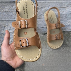 May include: Two brown leather sandals with buckle closures. The sandals have a light tan footbed and a stitched sole. The sandals are labeled "Mercan Barefoot Shoes".