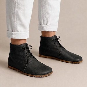 Puede incluir: Un par de botines negros con cordones negros finos y punta redondeada. Las botas cuentan con una suela plana y texturizada de color marrón con costuras visibles en el borde. Se llevan con pantalones blancos con dobladillo en los tobillos.