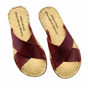 Peut inclure: Une paire de sandales à enfiler avec des brides croisées en cuir rouge foncé sur une semelle intérieure beige clair. La semelle porte l'inscription MerCan Barefoot Shoes et présente des coutures claires visibles sur le bord extérieur.
