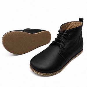 May include: A pair of black ankle boots featuring a smooth leather upper, black laces, and a tan-coloured sole with visible light-coloured stitching around the perimeter. One boot is upright, while the other is positioned to show its flat brown sole.