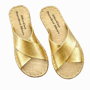Peut inclure: Une paire de sandales dorées métallisées avec des brides croisées sur le dessus du pied. La semelle intérieure est beige clair avec des coutures visibles sur le bord et le texte MerCan Barefoot Shoes imprimé en noir au niveau du talon.