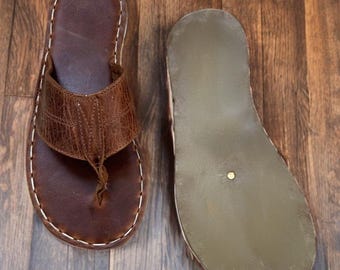 Può includere: Un paio di sandali infradito in pelle marrone. Un sandalo mostra il lato superiore con un cinturino in pelle testurizzata e cuciture bianche a contrasto lungo il bordo. L'altro sandalo è capovolto per mostrare la suola piatta in gomma verde oliva.