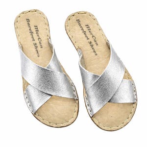 Op de afbeelding: Een paar zilveren leren sandalen met gekruiste bandjes. De sandalen hebben een lichtbruin voetbed en zijn aan de randen gestikt. De tekst "Mer-Can Barefoot Shoes" is op het voetbed gedrukt.