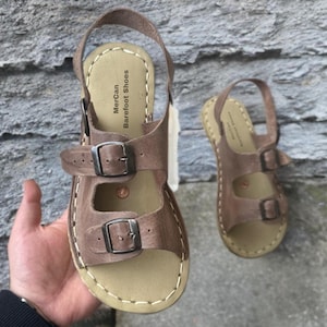Peut inclure: Une paire de sandales en cuir beige avec deux brides réglables dotées de boucles métalliques et une bride arrière. La semelle intérieure claire porte l'inscription MerCan Barefoot Shoes et un petit emblème circulaire cuivré.