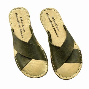 May include: A pair of olive green and tan leather sandals. The sandals feature a criss-cross strap design and are stitched with white thread. The text "MerCan Barefoot Shoes" is printed on the footbed.