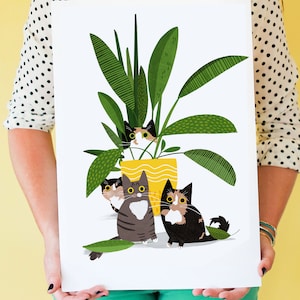 May include: A yellow planter with green leaves and four cats peeking out from behind the leaves. The cats are black, white, and gray with green eyes.