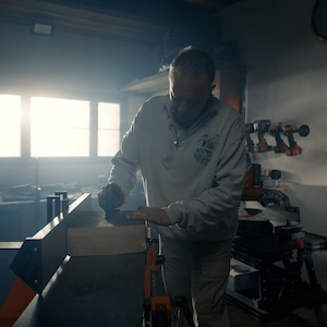 K&ouml;nnte beinhalten: Ein Mann in einem grauen Sweatshirt bedient eine Holzbearbeitungsmaschine und schleift ein St&uuml;ck Holz. Die Maschine hat orange und schwarze Akzente. Die Werkstatt ist schwach beleuchtet, mit einem hellen Fenster im Hintergrund.
