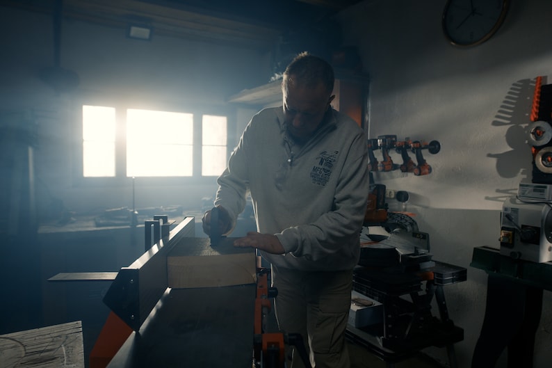 K&ouml;nnte beinhalten: Ein Mann in einem grauen Sweatshirt bedient eine Holzbearbeitungsmaschine und schleift ein St&uuml;ck Holz. Die Maschine hat orange und schwarze Akzente. Die Werkstatt ist schwach beleuchtet, mit einem hellen Fenster im Hintergrund.