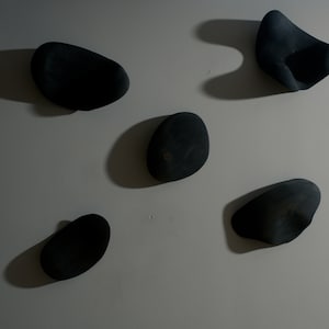 K&ouml;nnte beinhalten: F&uuml;nf schwarze, unregelm&auml;&szlig;ig geformte, glatte Steine sind an einer wei&szlig;en Wand befestigt. Die Steine sind in einem zuf&auml;lligen Muster angeordnet.