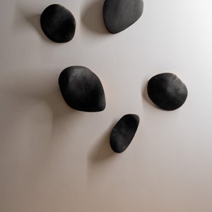 K&ouml;nnte beinhalten: Sechs schwarze, holzartige Wanddekorationen in Form unregelm&auml;&szlig;iger Steine, an einer wei&szlig;en Wand montiert.