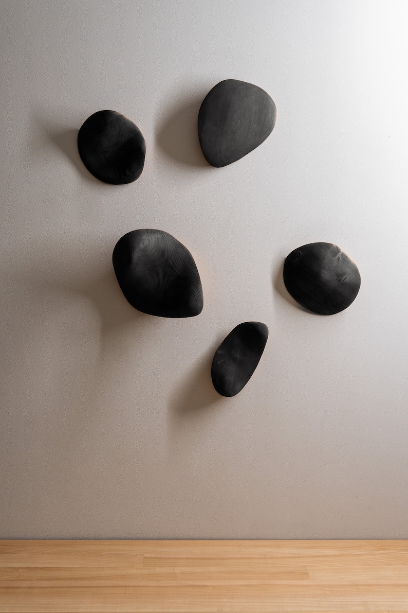 K&ouml;nnte beinhalten: Sechs schwarze, holzartige Wanddekorationen in Form unregelm&auml;&szlig;iger Steine, an einer wei&szlig;en Wand montiert.
