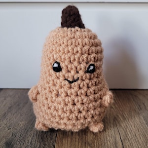 May include: A crocheted plush toy shaped like a pear with a brown stem and a smiling face. The pear is light brown and has black eyes and a mouth.