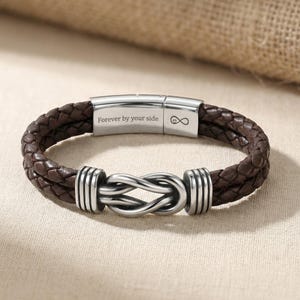 May include: A brown leather bracelet with a braided design. The bracelet features a silver-colored clasp with the inscription "Forever by your side" and an infinity symbol. The bracelet has a knot-shaped centerpiece.